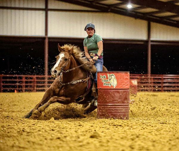 A rider and horse swiftly turning around a barrel in an indoor arena.