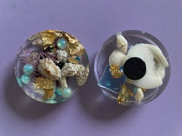 Two resin discs with embedded seashells, pearls, and gold flakes on a purple background.