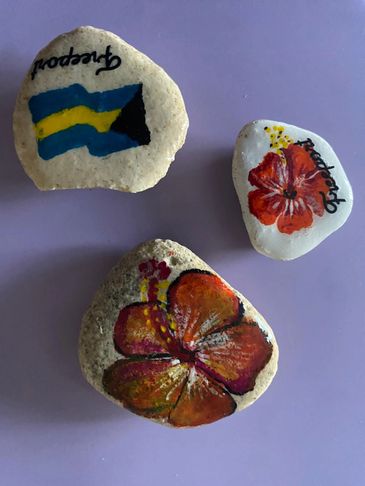 Three hand-painted rocks with flowers and a flag on a purple background.