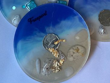 A round resin coaster with blue and beige ocean-like design and embedded seashells and gold flakes.
