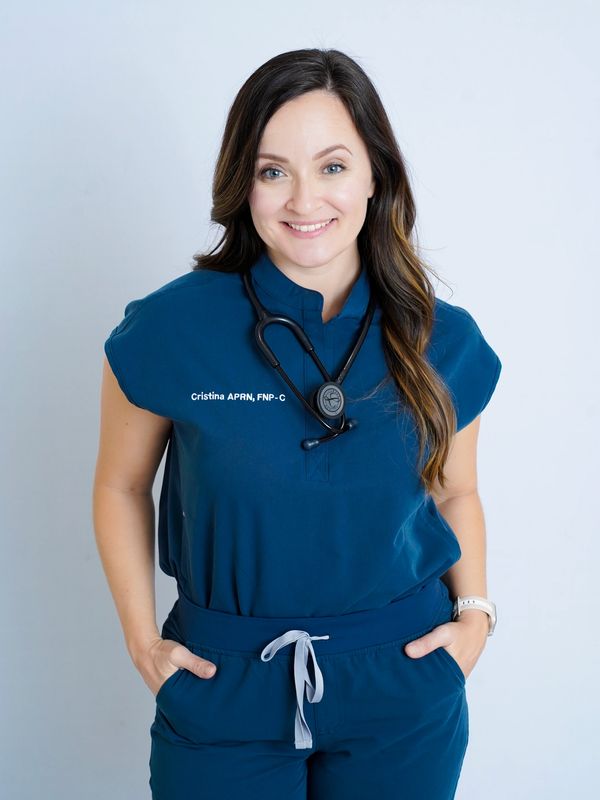 Smiling female nurse in blue scrubs with stethoscope and hands in pockets.
