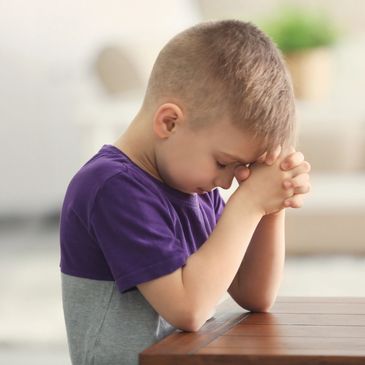 Young boy praying with closed eyes and clasped hands at a table.