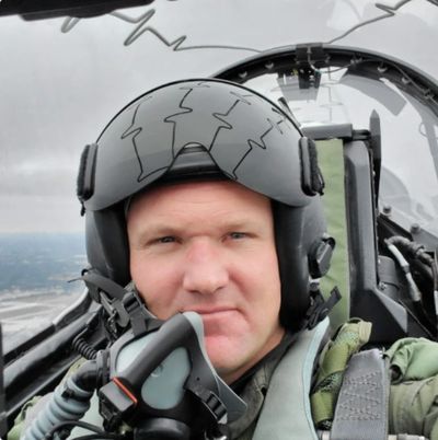 Pilot in a cockpit wearing a helmet and flight suit.