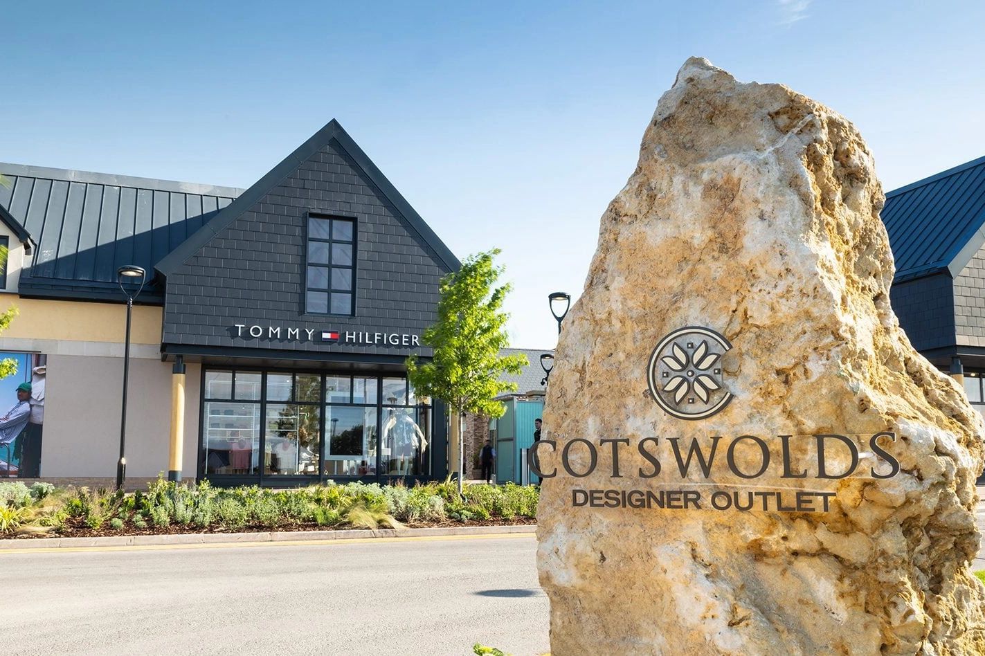 Large stone sign for Cotswolds Designer Outlet with Tommy Hilfiger store in the background.