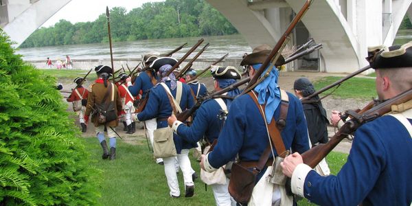Men in historical military uniforms march with muskets near a river under a bridge.