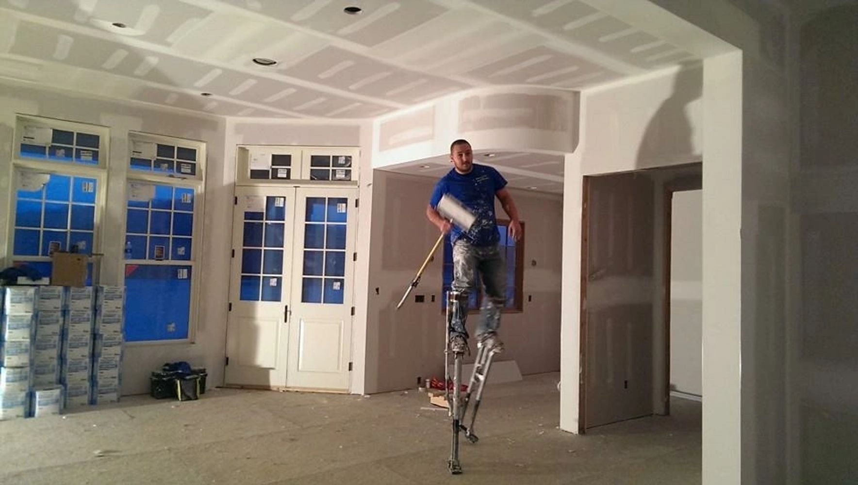 Worker on stilts applying drywall compound in a house under construction.