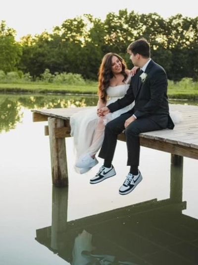 A bride and groom sitting on a wooden dock by a lake, smiling at each other.