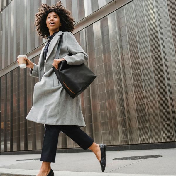 Woman in a gray coat walking confidently with coffee and a black bag.