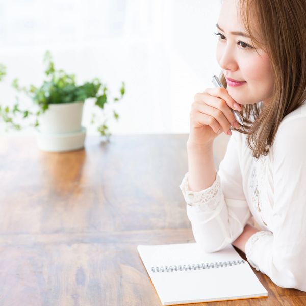 Woman in white shirt thoughtfully holding a pen near a notebook.