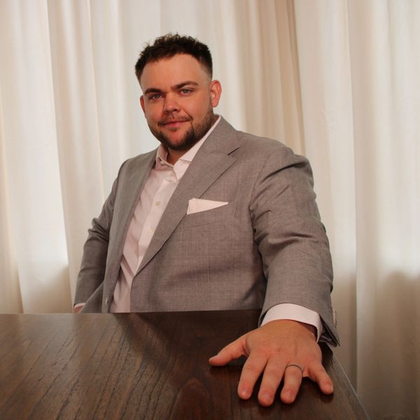 Man in a gray suit with a confident smile sitting at a wooden table.