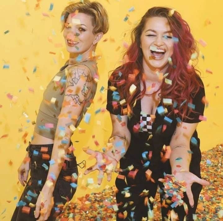 Two women joyfully throwing colorful confetti against a yellow backdrop.