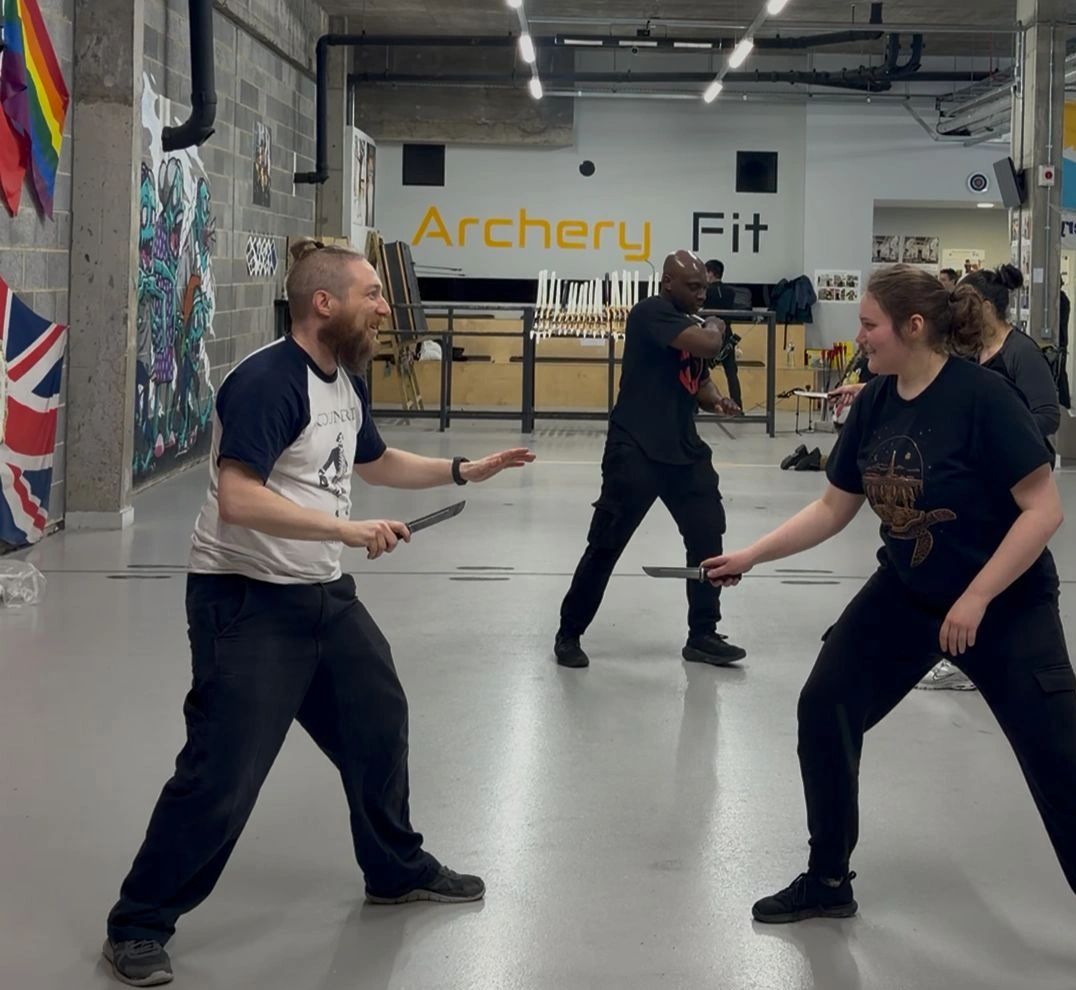 Two people practicing knife fighting in a gym with an instructor in the background.