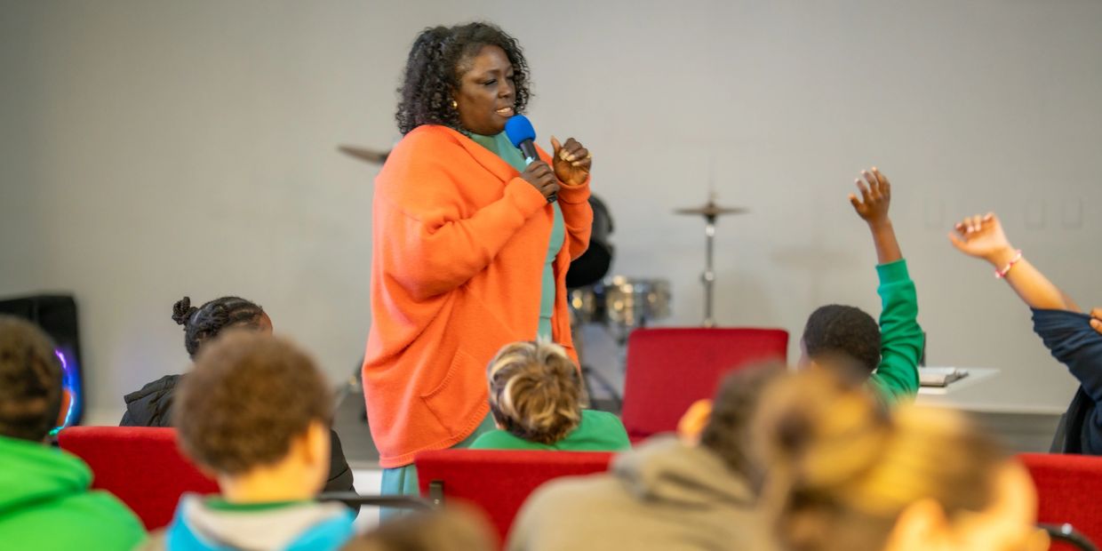 A woman speaking to a group of children, some raising their hands.