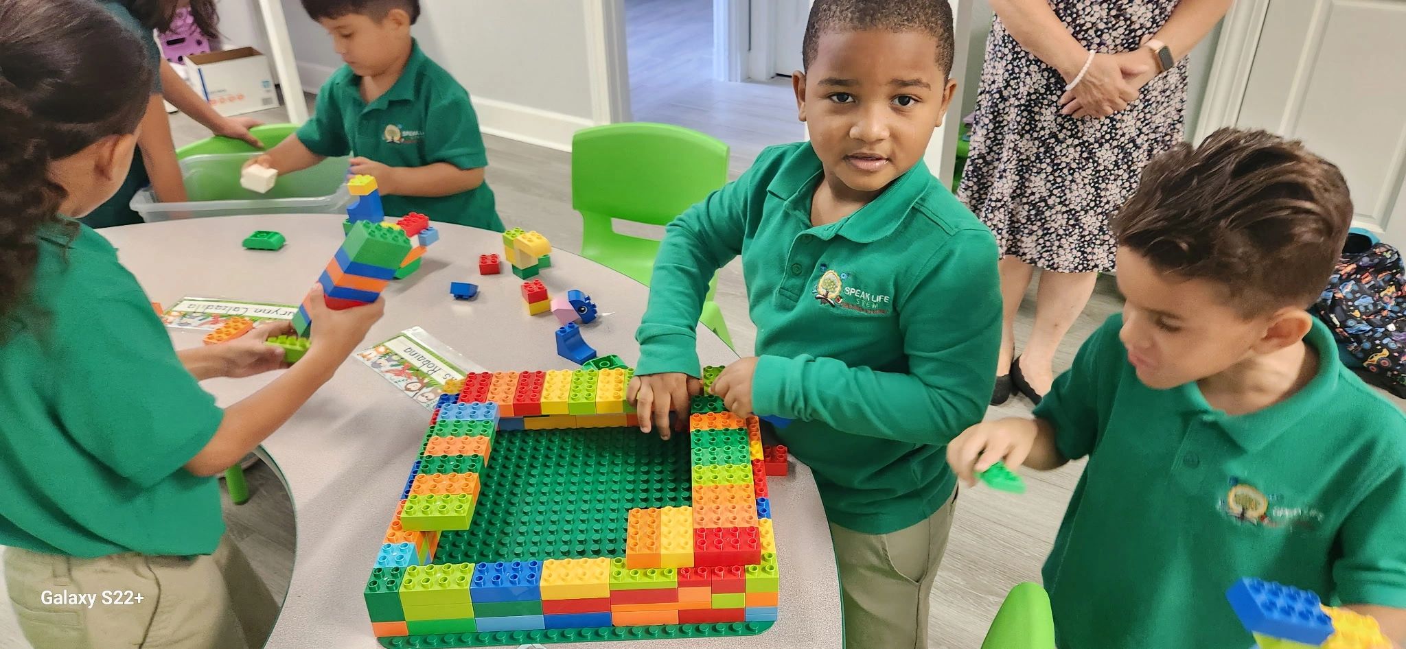 Children in green uniforms building with colorful Lego blocks in a classroom.