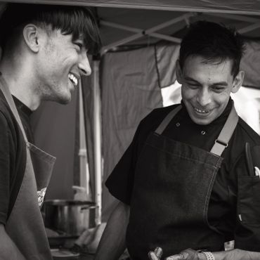 Two men smiling and wearing aprons, sharing a moment.