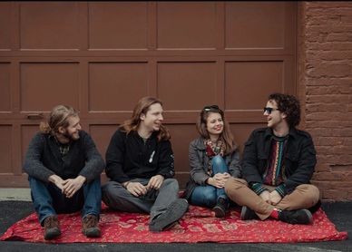 Four friends sitting on a red carpet, laughing and chatting outdoors.