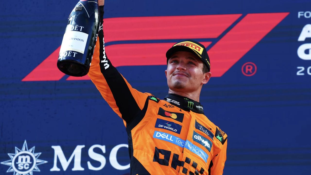 Lando Norris celebrates on the podium after winning the 2025 Austrian Grand Prix, holding a champagne bottle in McLaren team colours.