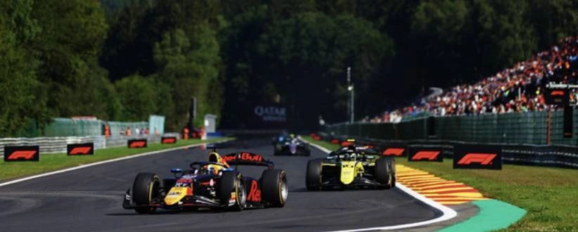 Formula 2 cars race side by side through Spa-Francorchamps with spectators watching from packed grandstands on a sunny race day. Formula 2 cars race side by side through Spa-Francorchamps with spectators watching from packed grandstands on a sunny race day.