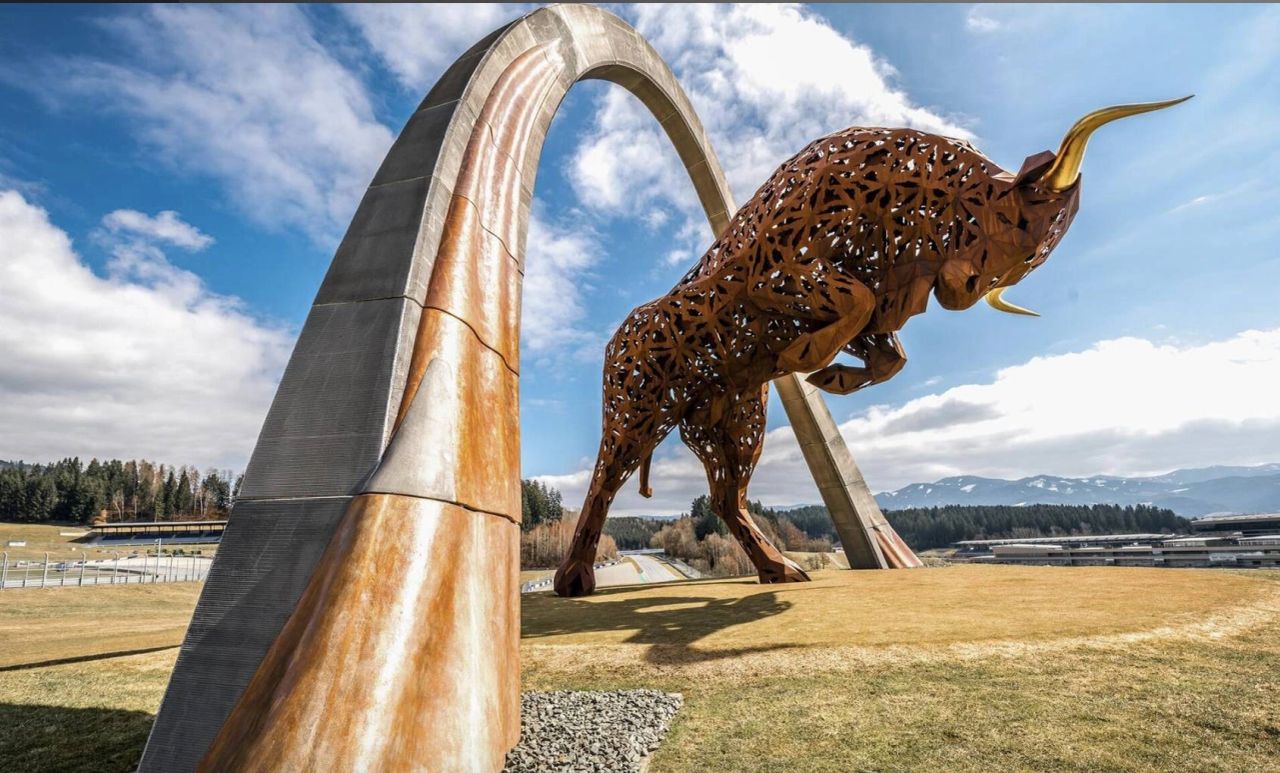 Large bronze bull statue under a metallic arch at the Red Bull Ring circuit in Spielberg, Austria, with Styrian mountains in the background. Large bronze bull statue under a metallic arch at the Red Bull Ring circuit in Spielberg, Austria, with Styrian mountains in the background.