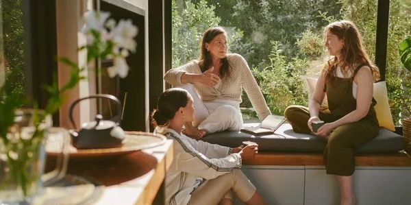Three women having a conversation indoors by a window.