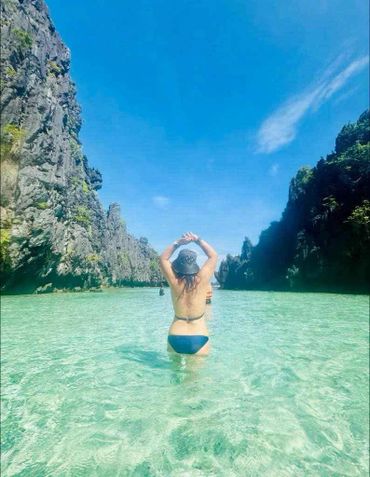 Woman in blue bikini standing in clear water between rocky cliffs under a bright blue sky.