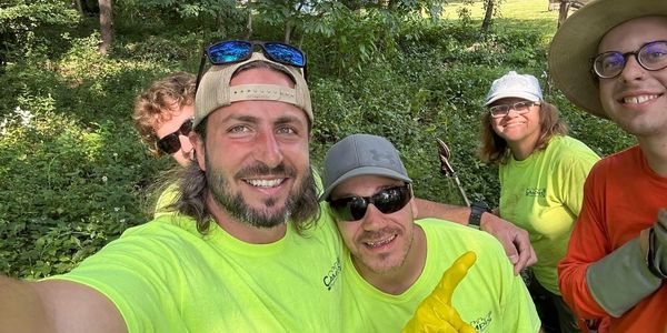 A group of five smiling friends in bright outdoor work attire taking a selfie in a green wooded area.