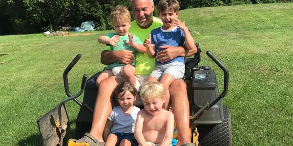 Man and four kids enjoying a ride on a lawn mower in a sunny yard.