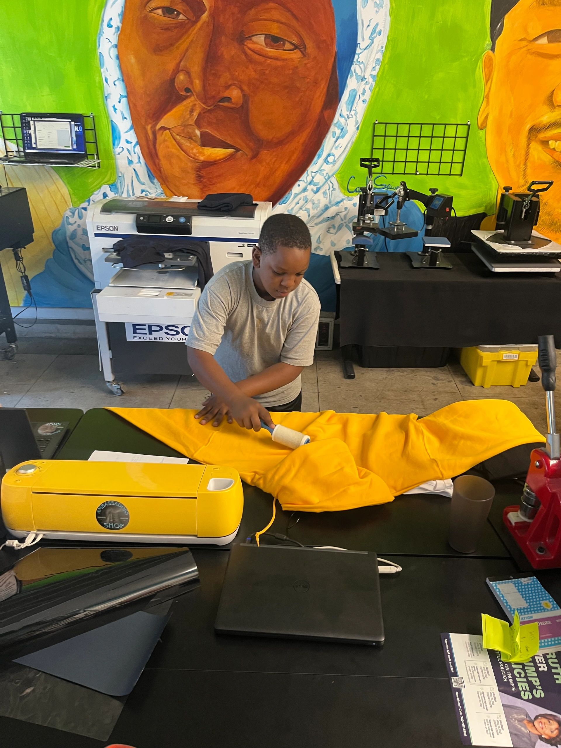 A boy uses a lint roller on a yellow hoodie in a colorful workshop.