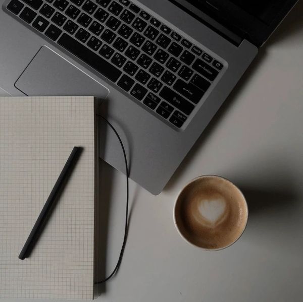 A laptop, a notebook with a pen, and a cup of coffee with heart-shaped foam on a table.
