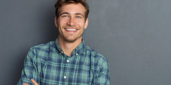 Smiling young man in a green plaid shirt stands against a gray background.