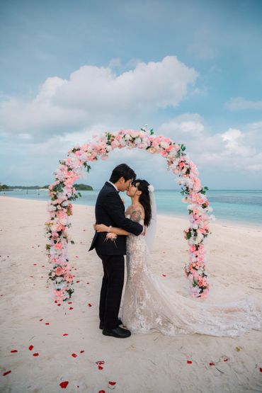 A couple kissing under a floral arch on a beach during their wedding.