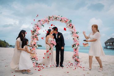 A couple kisses under a floral arch on a beach while flower petals are thrown.