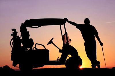 Silhouette of a golfer leaning on a golf cart at sunset.
