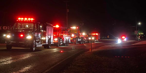 A line of fire trucks with flashing red lights on a dark road at night.