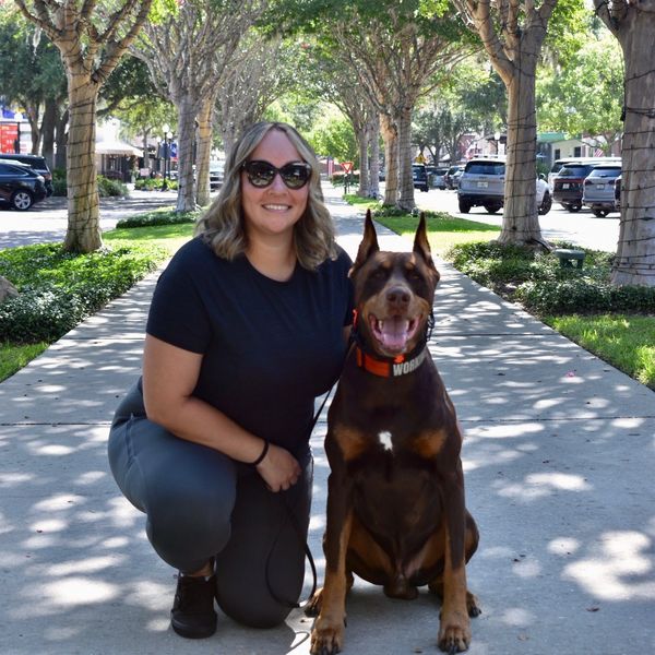 Woman kneeling next to a happy Doberman on a shaded sidewalk.