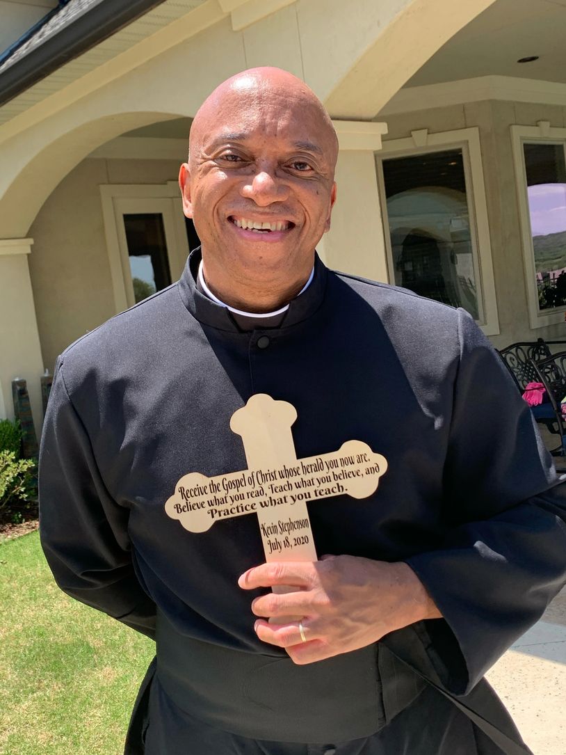 A smiling priest in black robe holding a wooden cross with an inspirational quote.