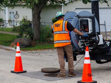 Worker operating machinery by an open manhole with safety cones.