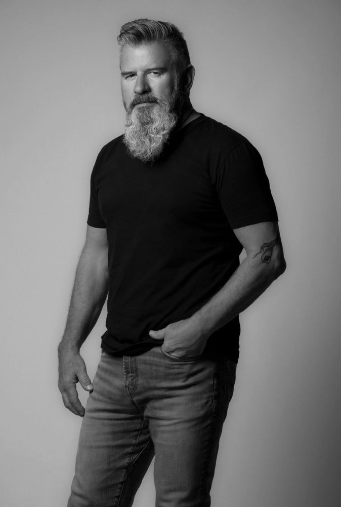 A man wearing a black T-shirt and jeans and a long beard is seen in the front.