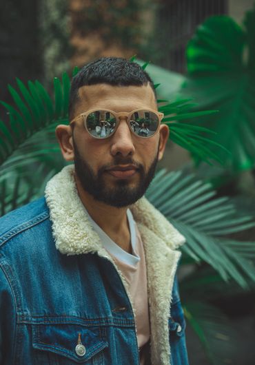 Man wearing sunglasses and a denim jacket with a shearling collar, surrounded by green leaves.