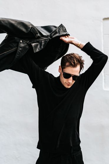 Man wearing sunglasses putting on a black leather jacket against a white wall.