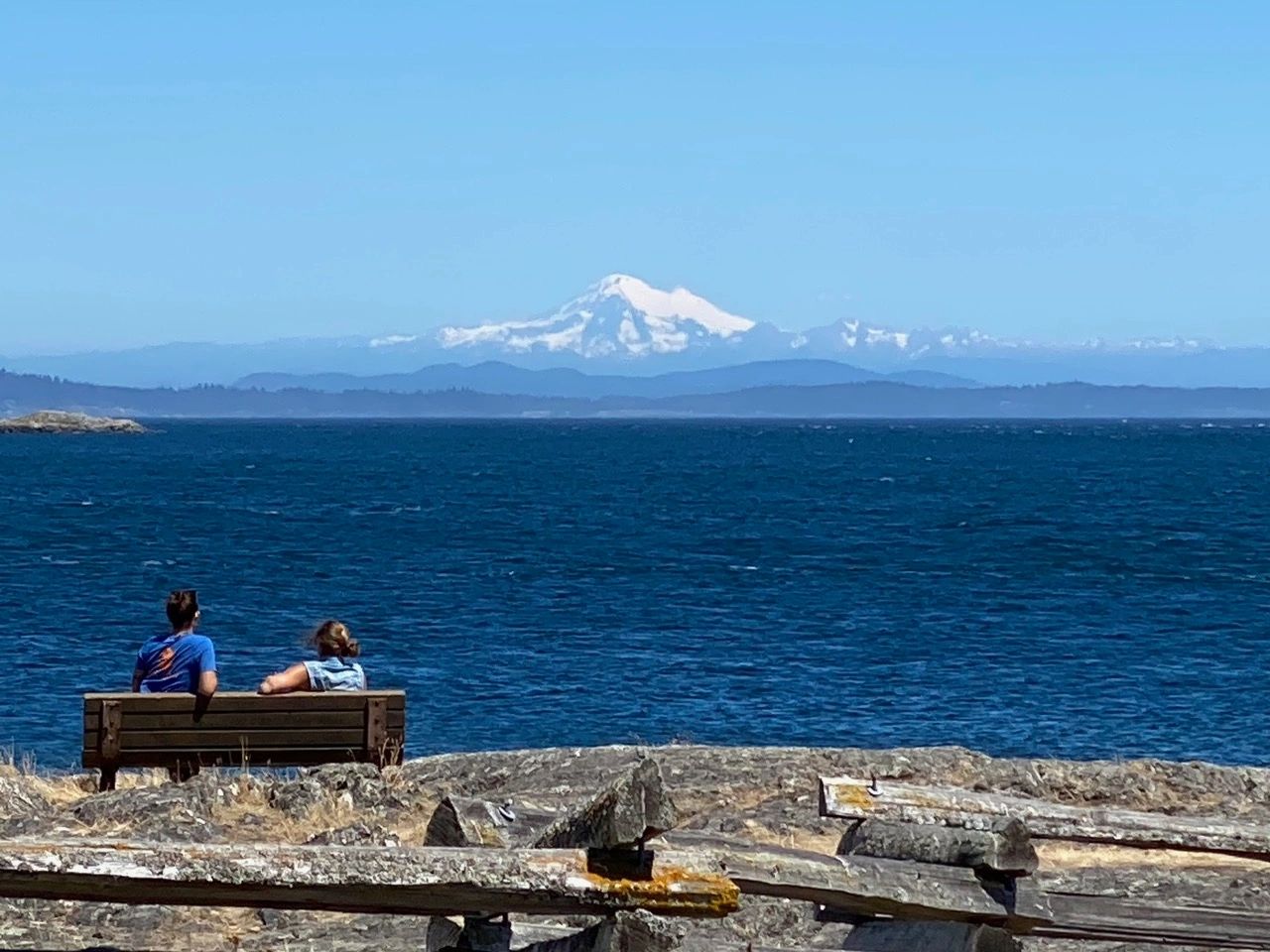 Two people sitting on a bench looking to the Ocean and Mountains