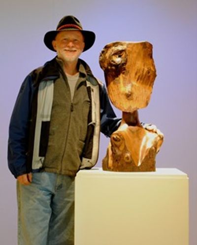 Man in hat and jacket next to a wooden bust sculpture on a white pedestal