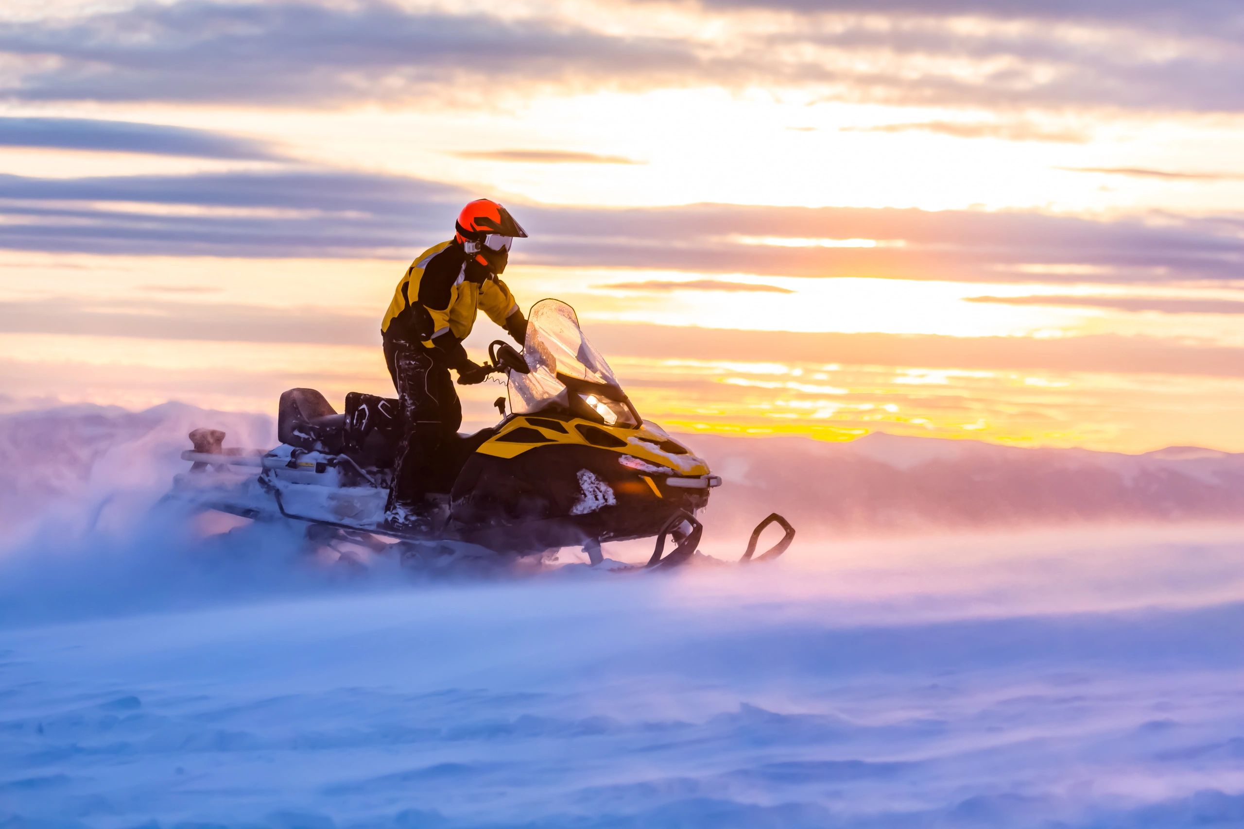 A man riding a snowmobile across a snowy landscape at sunset
