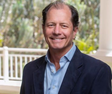 Smiling man in a blue shirt and dark blazer standing outdoors.