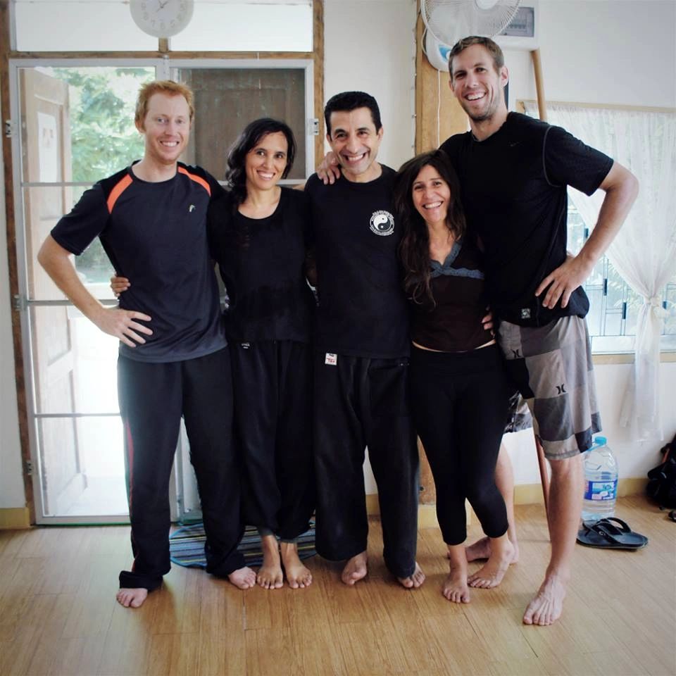 Five smiling people standing barefoot indoors, arms around each other.