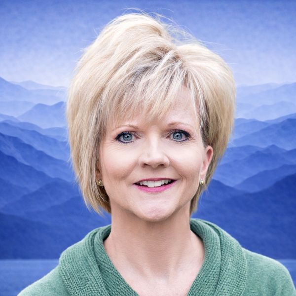 Smiling woman with blonde hair and blue eyes in front of a blue mountain backdrop.