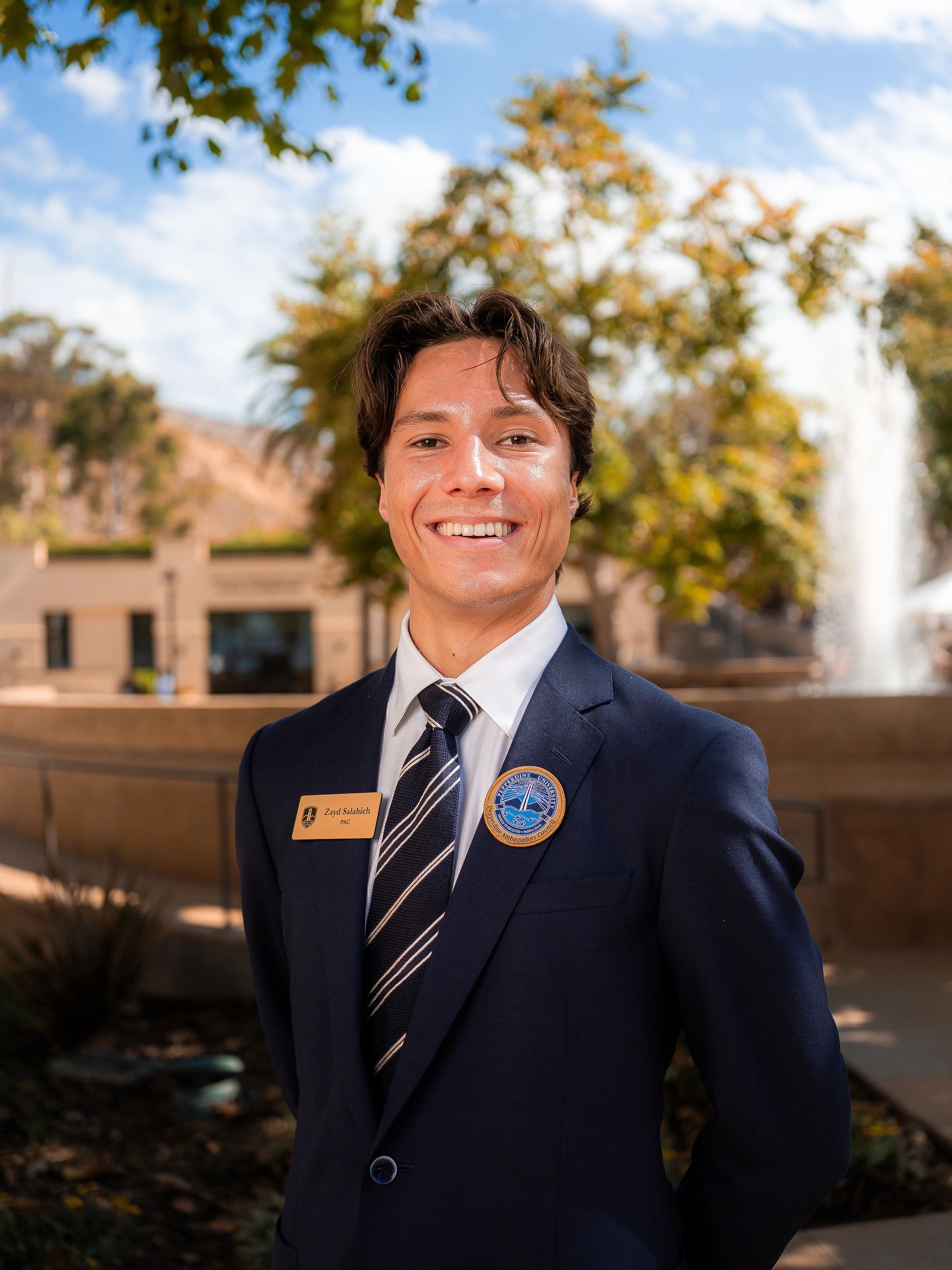 Young man in a suit smiling outdoors on a sunny day.