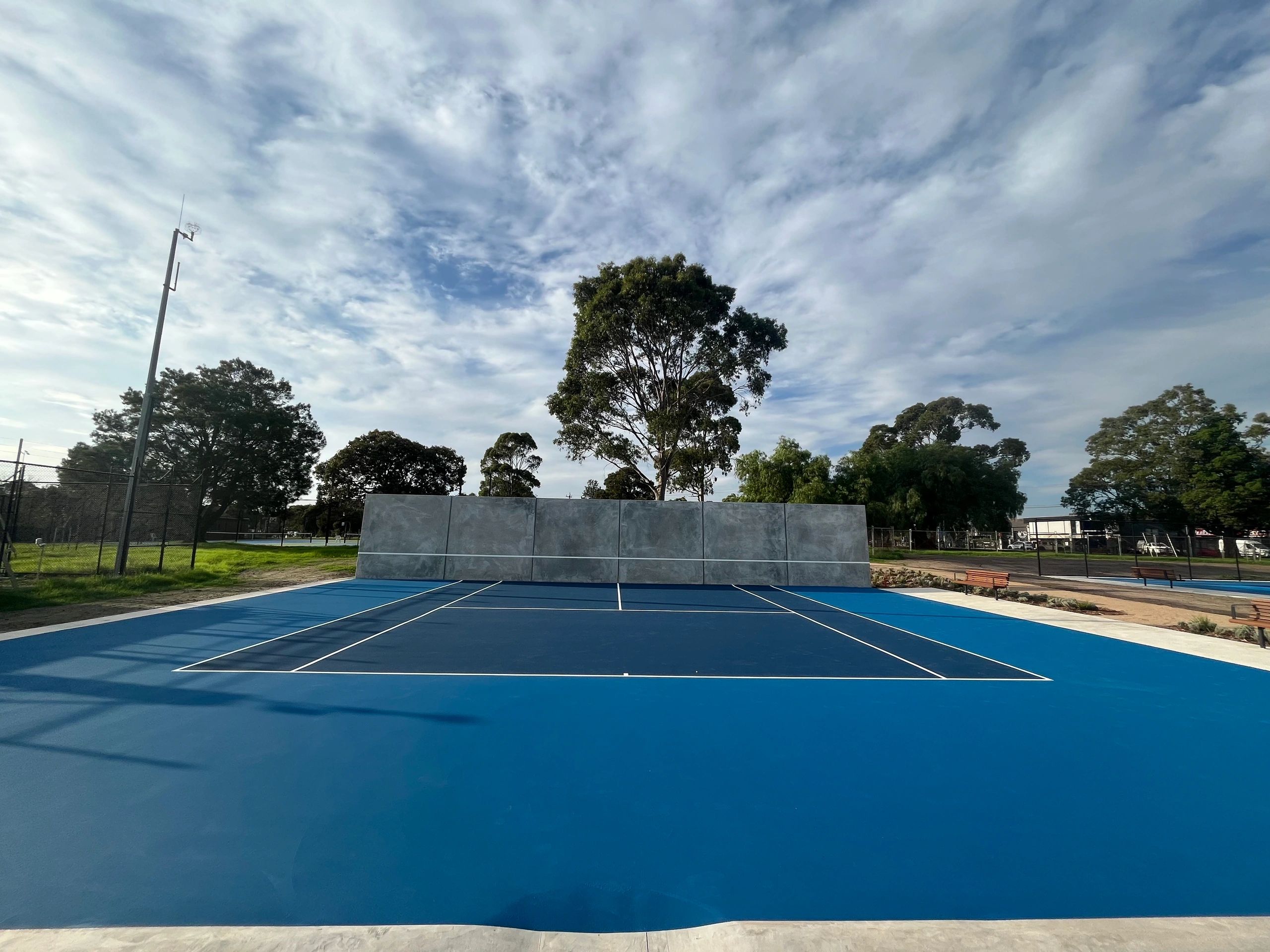 Blue sports court with a tall concrete wall against a backdrop of trees and a cloudy sky.