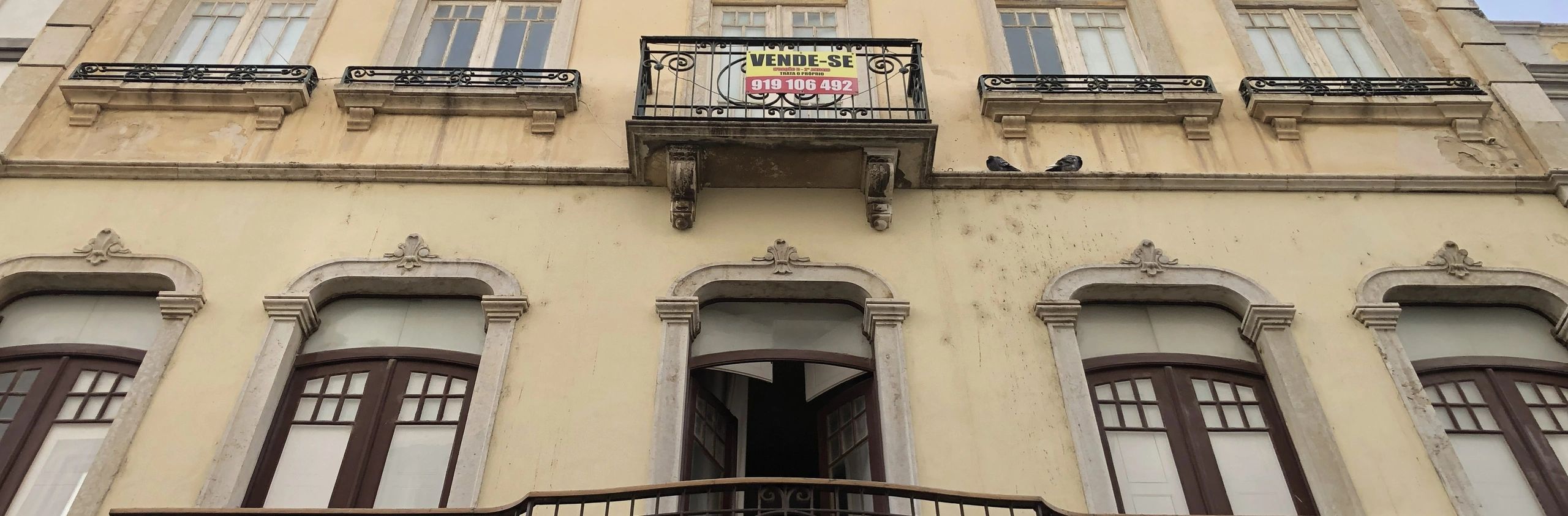 Old building facade with a 'For Sale' sign and pigeons on the balcony.
