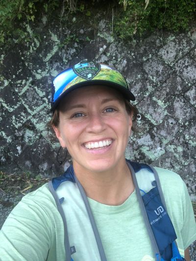 A smiling woman outdoors wearing a cap and hydration pack.